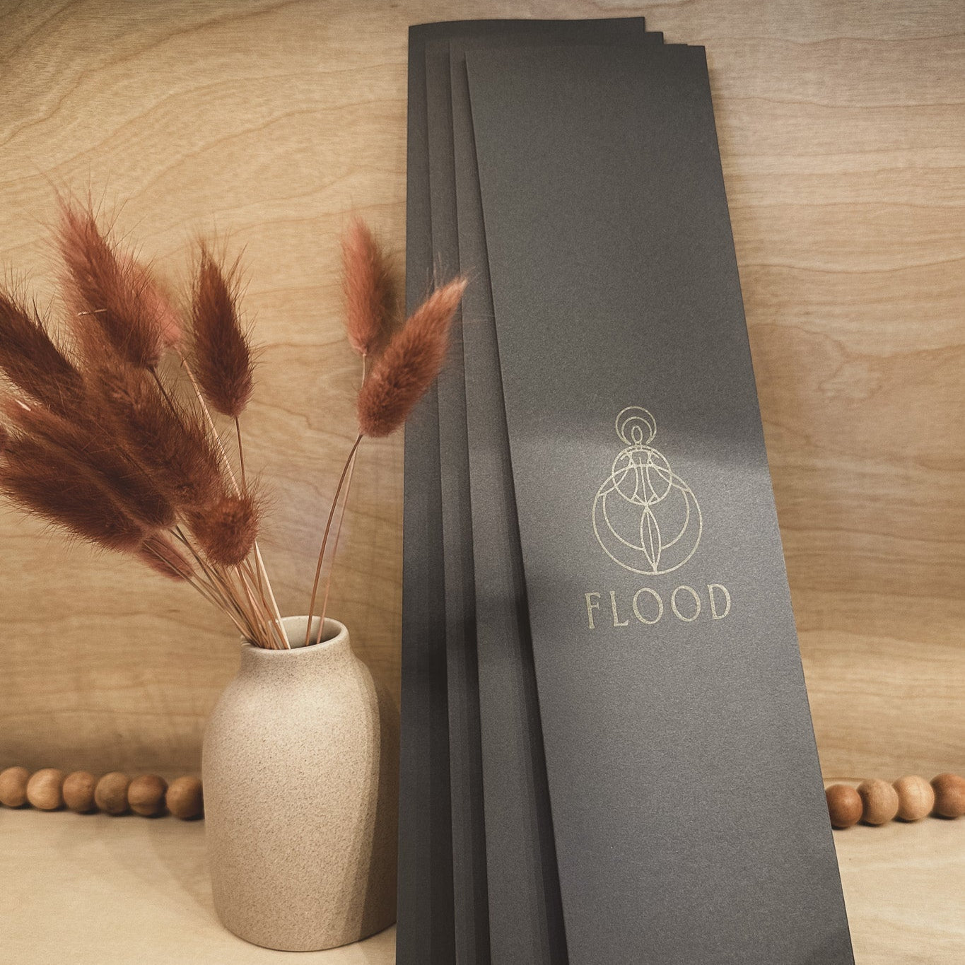 Dark gray booklets with 'FLOOD' branding on a wooden surface with a vase and pampas grass.