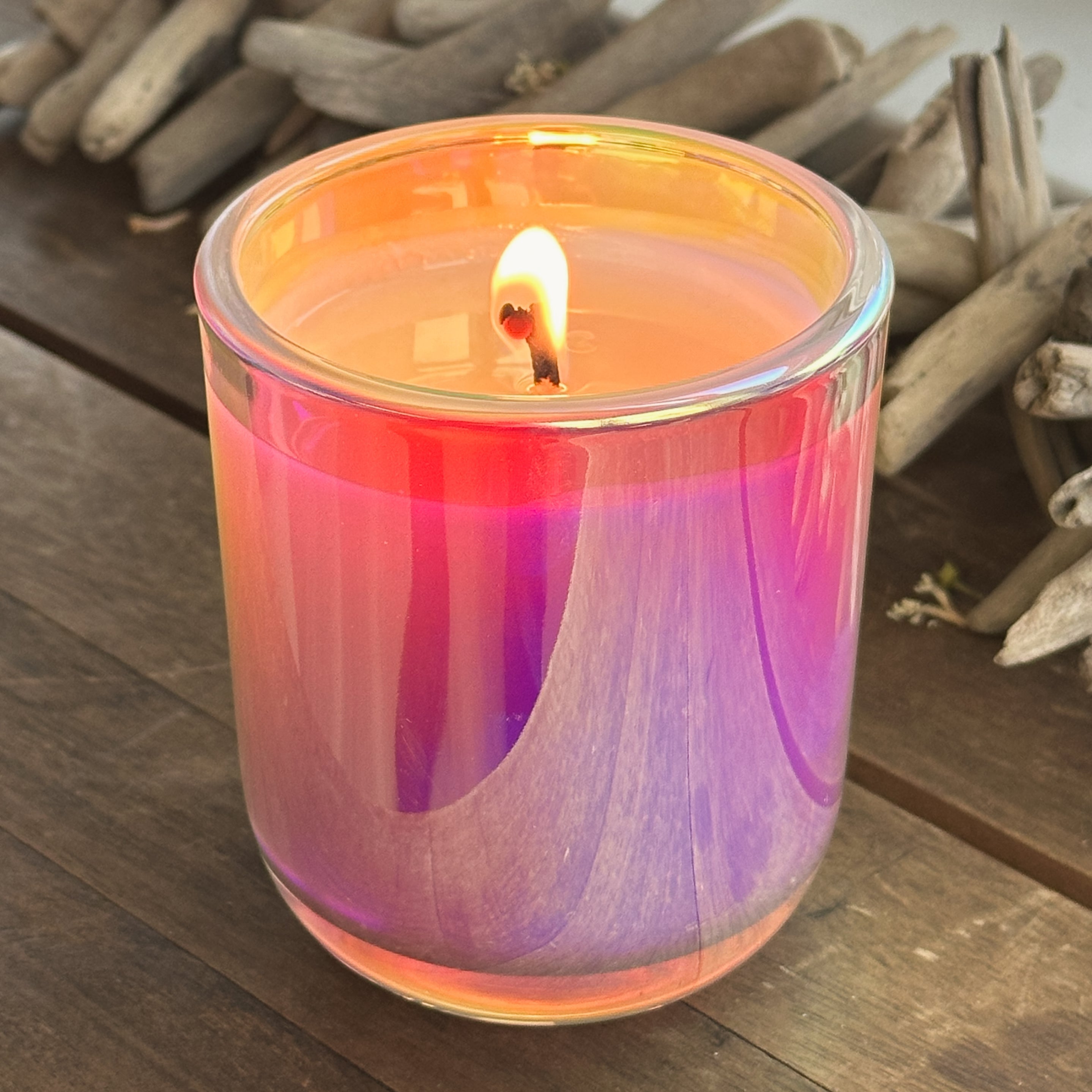 Candle in a glass jar with a colorful design on a wooden surface with driftwood in the background.